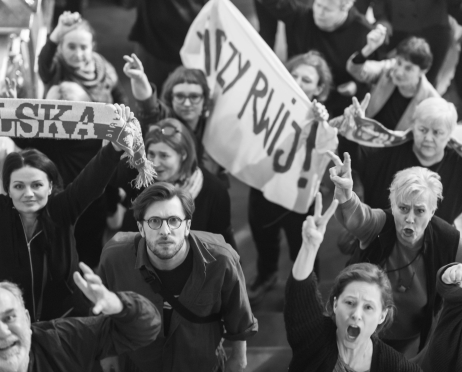 Czarno-białe zdjęcie. Grupa protestujących widzianych z góry. Uczestnicy trzymają transparenty i szaliki, a część z nich unosi dłonie w geście sprzeciwu lub skanduje hasła.